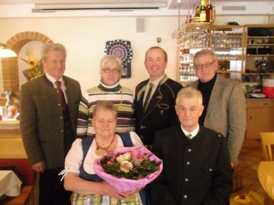 Eine Gruppe von sechs Personen steht in einem Restaurant, wobei eine Frau in der Mitte einen Blumenstrauß hält. Hinter ihnen befindet sich ein Dartboard und Regale mit Gläsern.