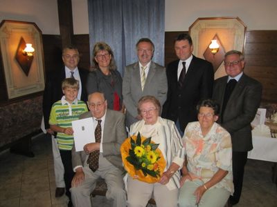 Eine Gruppe von Menschen, darunter ein älteres Paar, posiert für ein Foto in einem Raum mit einem Tisch und Blumen. Das ältere Paar sitzt auf einer Bank.
