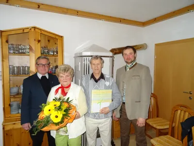 Vier Personen stehen in einem Raum, einer hält einen Blumenstrauß, ein anderer ein verpacktes Geschenk. Der Mann rechts trägt eine Anzugjacke.