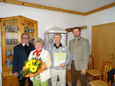 Vier Personen stehen in einem Raum, einer hält einen Blumenstrauß, ein anderer ein verpacktes Geschenk. Der Mann rechts trägt eine Anzugjacke.