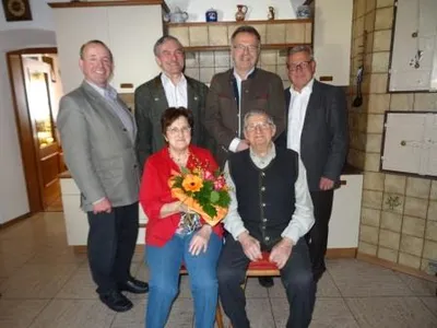 Fünf Personen posieren für ein Foto in einem Raum. Ein älterer Mann sitzt auf einem Stuhl und eine Frau in einer roten Jacke hält einen Blumenstrauß. Dahinter stehen drei Männer in formeller Kleidung.
