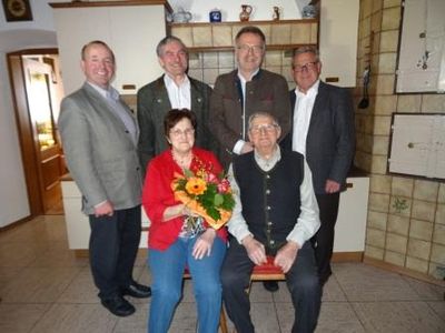 Fünf Personen posieren für ein Foto in einem Raum. Ein älterer Mann sitzt auf einem Stuhl und eine Frau in einer roten Jacke hält einen Blumenstrauß. Dahinter stehen drei Männer in formeller Kleidung.