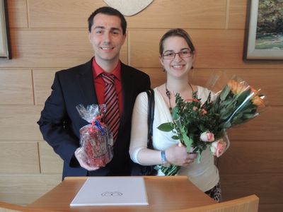 Ein Mann und eine Frau stehen nebeneinander, lächelnd, wobei der Mann ein verpacktes Geschenk und die Frau einen Blumenstrauß hält. Sie stehen vor einer Holzwand mit einem gerahmten Bild.