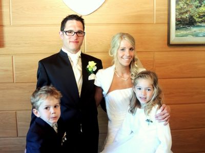 Ein Mann und eine Frau in formeller Kleidung, lächelnd, stehen mit zwei Kindern vor einer Holzwand. Der Mann trägt eine Brille und eine Ansteckblume, und die Frau trägt eine Halskette. Die Kinder tragen weiße Kleider.