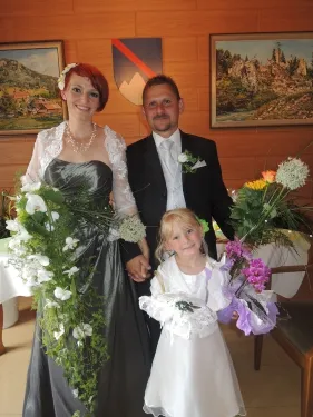 Eine Braut in einem grünen Kleid und ein Bräutigam in einem schwarzen Anzug stehen mit einem kleinen Mädchen, das Blumen hält, vor einer Wand mit gerahmten Bildern.