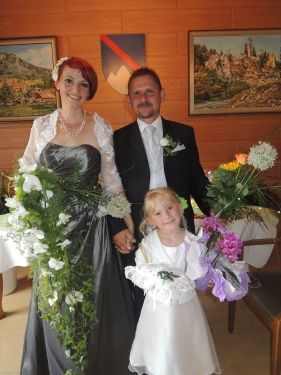 Eine Braut in einem grünen Kleid und ein Bräutigam in einem schwarzen Anzug stehen mit einem kleinen Mädchen, das Blumen hält, vor einer Wand mit gerahmten Bildern.