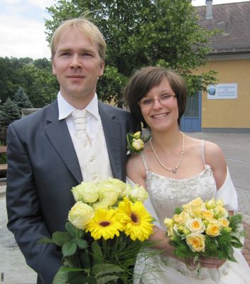 Ein frisch verheiratetes Paar steht draußen, der Bräutigam in einem Anzug und die Braut in einem weißen Hochzeitskleid, beide halten Blumensträuße. Hinter ihnen steht ein Gebäude mit einem Schild und Bäumen.
