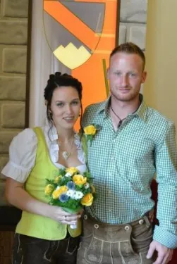 Ein Mann und eine Frau in traditioneller deutscher Kleidung stehen nah beieinander und lächeln für ein Foto. Die Frau hält einen Strauß gelber und weißer Blumen. Hinter ihnen ist eine dekorative Wand mit einem Schilddesign.
