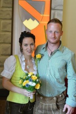 Ein Mann und eine Frau in traditioneller deutscher Kleidung stehen nah beieinander und lächeln für ein Foto. Die Frau hält einen Strauß gelber und weißer Blumen. Hinter ihnen ist eine dekorative Wand mit einem Schilddesign.