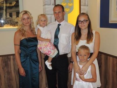 Eine Familie aus vier Personen steht in einem Raum zusammen, mit zwei Frauen, einem Mann und zwei kleinen Mädchen. Der Mann trägt eine Krawatte, und die Frauen sind in Kleidern. Die Mädchen halten die Hände des Mannes.