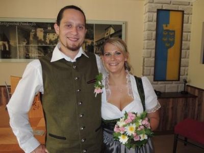 Ein Mann und eine Frau in traditioneller deutscher Kleidung stehen lächelnd nebeneinander, der Mann hält einen Blumenstrauß. Sie befinden sich in einem Raum mit einer Ziegelwand und einem gerahmten Bild dahinter.