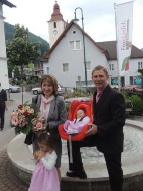 Eine Familie steht vor einem Brunnen mit einem Baby im Kinderwagen, eine Frau hält Blumen und ein Mann in Anzug. Hinter ihnen ist eine Kirche und ein Banner zu sehen.