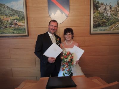 Ein frisch verheiratetes Paar steht vor einer Holzwand, hält ihre Heiratsurkunden und lächelt. Die Braut trägt ein Hochzeitskleid und hält einen Blumenstrauß. Hinter ihnen hängen zwei gerahmte Gemälde an der Wand.
