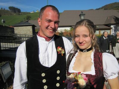 Ein Mann und eine Frau in traditioneller deutscher Kleidung, lächeln und posieren für ein Foto vor einem Haus mit einem grünen Hügel im Hintergrund.