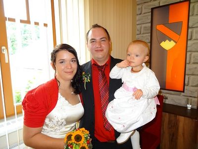 Ein Mann und eine Frau in Hochzeitskleidung posieren mit einem Baby vor einer Ziegelwand mit einem Gemälde.