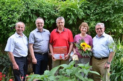 Fünf Erwachsene stehen lächelnd in einem Garten. Ein Mann in Rot hält eine Geschenkbox und eine Frau in Pink hält Blumen.