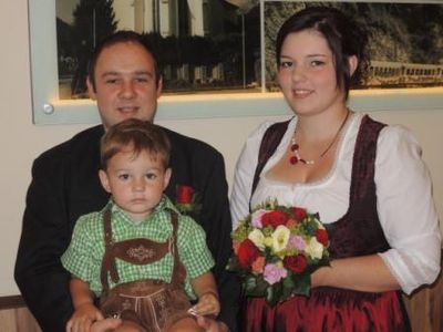 Ein Mann und eine Frau in traditioneller deutscher Kleidung, die einen Blumenstrauß halten, posieren für ein Foto mit einem jungen Jungen in traditioneller bayerischer Kleidung.