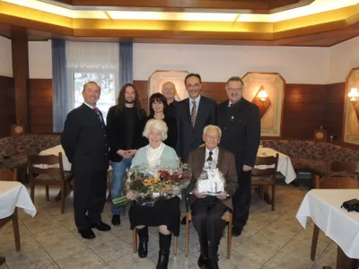 Eine Gruppe von Menschen in formeller Kleidung steht und sitzt in einem Restaurant, wobei eine ältere Frau Blumen hält und ein Mann ein Geschenk in der Hand hat.