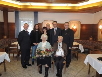Eine Gruppe von Menschen in formeller Kleidung steht und sitzt in einem Restaurant, wobei eine ältere Frau Blumen hält und ein Mann ein Geschenk in der Hand hat.