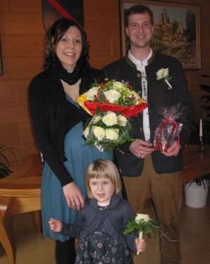 Eine Familie aus drei Personen, bestehend aus einer schwangeren Frau, einem Mann und einem kleinen Mädchen, steht zusammen, hält Blumen und ein Geschenk und lächelt für ein Foto in einem Raum.