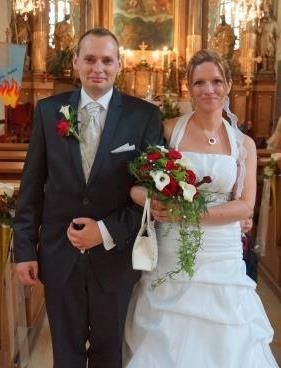 Ein frisch verheiratetes Paar steht in einem Kirchenschiff, der Bräutigam in einem schwarzen Anzug und die Braut in einem weißen Kleid, das einen Blumenstrauß hält.
