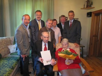 Eine Gruppe von Menschen in formeller Kleidung versammelt sich um ein älteres Paar. Der ältere Mann hält ein Zertifikat, und die ältere Frau hält einen Blumenstrauß. Sie befinden sich in einem Wohnzimmer mit einer Couch und einer Holztür.