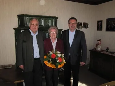 Drei Personen stehen in einem Raum mit einem Blumenstrauß in der Mitte. Sie sind formell gekleidet. Hinter ihnen steht ein Holzschrank und ein Tisch mit einem dekorativen Gegenstand.