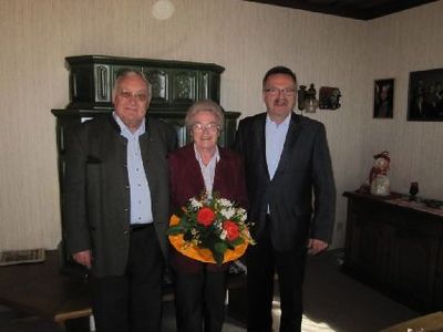 Drei Personen stehen in einem Raum mit einem Blumenstrauß in der Mitte. Sie sind formell gekleidet. Hinter ihnen steht ein Holzschrank und ein Tisch mit einem dekorativen Gegenstand.