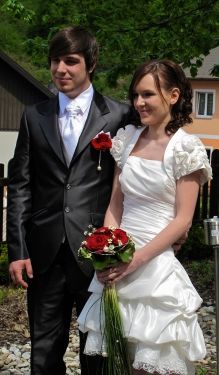 Ein frisch verheiratetes Paar steht draußen, der Bräutigam in einem schwarzen Anzug und die Braut in einem weißen Hochzeitskleid, das einen Strauß roter Rosen hält. Sie sind von Kies und einem Holzzaun umgeben.