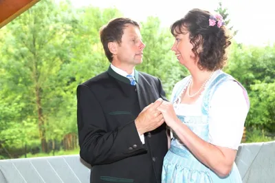 Ein Mann und eine Frau in traditioneller Kleidung stehen zusammen, lächeln und halten Händchen. Sie befinden sich auf einem Balkon mit Bäumen im Hintergrund.