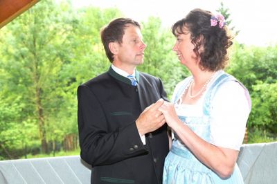 Ein Mann und eine Frau in traditioneller Kleidung stehen zusammen, lächeln und halten Händchen. Sie befinden sich auf einem Balkon mit Bäumen im Hintergrund.