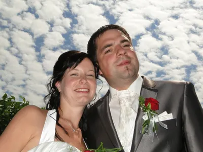 Ein frisch verheiratetes Paar, der Bräutigam in einem grauen Anzug mit einer roten Rose und die Braut in einem weißen Kleid, lächeln unter einem blauen Himmel mit Wolken.