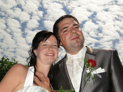 Ein frisch verheiratetes Paar, der Bräutigam in einem grauen Anzug mit einer roten Rose und die Braut in einem weißen Kleid, lächeln unter einem blauen Himmel mit Wolken.