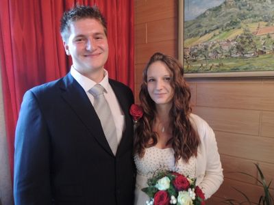 Ein Mann und eine Frau stehen eng beieinander und lächeln für ein Foto. Der Mann trägt einen Anzug und eine Krawatte, während die Frau einen Blumenstrauß hält. Hinter ihnen ist ein roter Vorhang und ein gerahmtes Landschaftsgemälde.