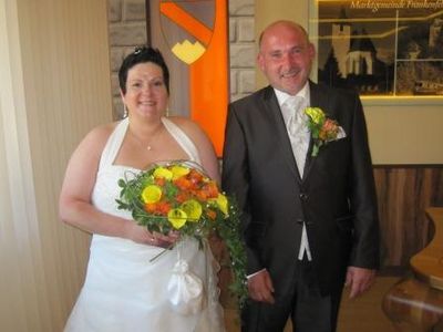 Eine Braut in einem weißen Kleid, das einen Blumenstrauß hält, steht neben einem Bräutigam in einem schwarzen Anzug, beide lächeln in die Kamera.
