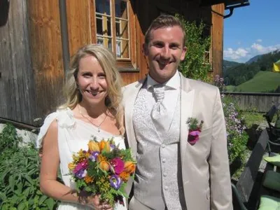 Ein frisch verheiratetes Paar steht vor einem rustikalen Holzhaus, die Braut hält einen bunten Blumenstrauß und der Bräutigam lächelt in die Kamera.