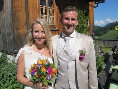 Ein frisch verheiratetes Paar steht vor einem rustikalen Holzhaus, die Braut hält einen bunten Blumenstrauß und der Bräutigam lächelt in die Kamera.