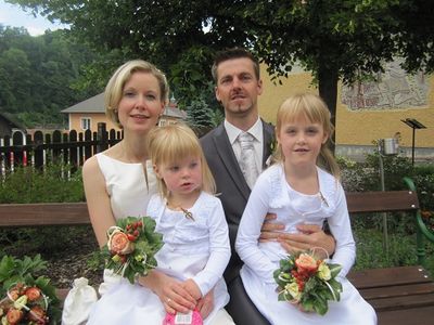 Ein frisch verheiratetes Paar und ihre beiden kleinen Töchter posieren für ein Foto auf einer Parkbank. Das Paar ist feierlich gekleidet, und die Mädchen halten Blumensträuße.