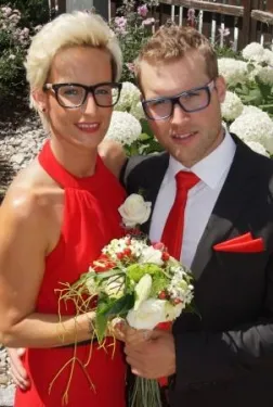 Ein Mann und eine Frau in formeller Kleidung halten einen Blumenstrauß und stehen eng nebeneinander in einer Gartenumgebung.