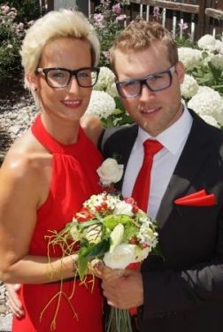 Ein Mann und eine Frau in formeller Kleidung halten einen Blumenstrauß und stehen eng nebeneinander in einer Gartenumgebung.