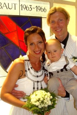 Eine Familie aus drei Personen, ein Mann, eine Frau und ein Baby, lächelt für ein Foto. Die Frau hält einen Strauß weißer Blumen. Der Mann trägt einen Anzug und eine Krawatte. Hinter ihnen befindet sich ein Buntglasfenster mit der Aufschrift 'BAU 1963-1966'.
