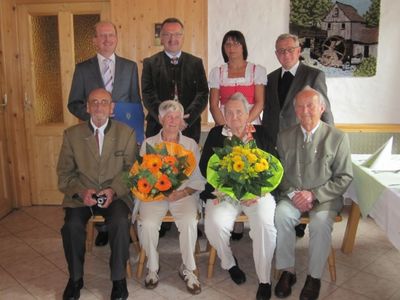 Eine Gruppe von Menschen in formeller Kleidung sitzt auf einer Bank und hält Blumen, ein lächelnder Mann hält eine Kamera im Vordergrund. Hinter ihnen steht eine Frau in traditioneller Kleidung neben einem Mann in Anzug. Der Hintergrund zeigt eine Holzwand mit einem Bilderrahmen.