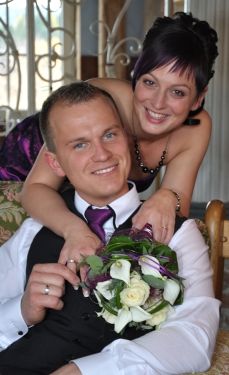 Ein Mann und eine Frau sitzen auf einer Couch, die Frau hält einen Blumenstrauß und lächelt, während der Mann in die Kamera lächelt.