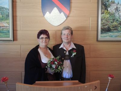 Ein Mann und eine Frau stehen lächelnd eng nebeneinander vor einer Holzwand mit gerahmten Bildern und einem Wappen. Die Frau hält einen Blumenstrauß.
