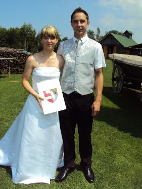 Ein frisch verheiratetes Paar steht auf einem Rasenfeld, die Frau in einem weißen Hochzeitskleid hält ein gerahmtes Zertifikat, der Mann in formeller Kleidung, mit einem alten Wagen und einer rustikalen Scheune im Hintergrund.
