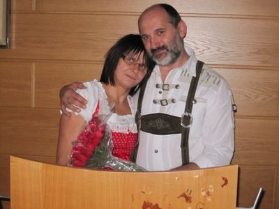 Ein Mann und eine Frau in traditioneller deutscher Kleidung stehen eng beieinander und lächeln, wobei der Mann seinen Arm um die Schulter der Frau gelegt hat. Sie stehen vor einer Holzwand.