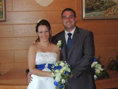 Ein frisch verheiratetes Paar steht eng beieinander und lächelt für ein Foto vor einer Holzwand mit gerahmten Bildern. Die Frau hält einen Strauß aus blauen und weißen Blumen.