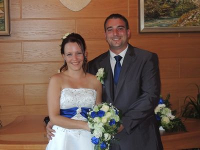 Ein frisch verheiratetes Paar steht eng beieinander und lächelt für ein Foto vor einer Holzwand mit gerahmten Bildern. Die Frau hält einen Strauß aus blauen und weißen Blumen.