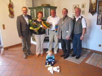 Fünf Erwachsene und ein Kleinkind stehen in einem Raum mit gefliestem Boden, eine Frau hält Blumen und ein Baby sitzt auf dem Boden mit Spielzeug.