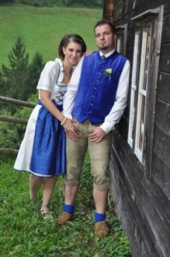 Ein Mann und eine Frau in traditioneller bayerischer Kleidung stehen neben einem Holzhaus mit einem grünen Feld im Hintergrund.